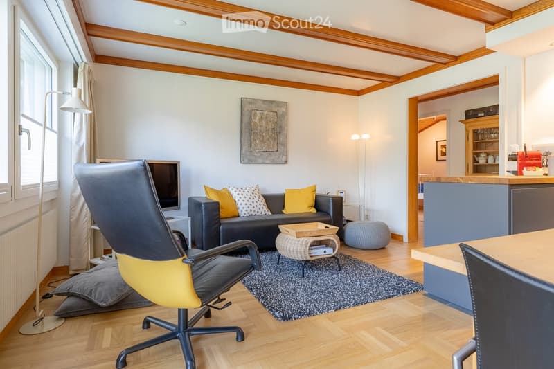 Bright living room with wooden floors, exposed wooden beams, large windows, a black leather sofa with yellow and white pillows, a modern chair, a coffee table, and a small ottoman. A floor lamp and a piece of abstract art are also visible.