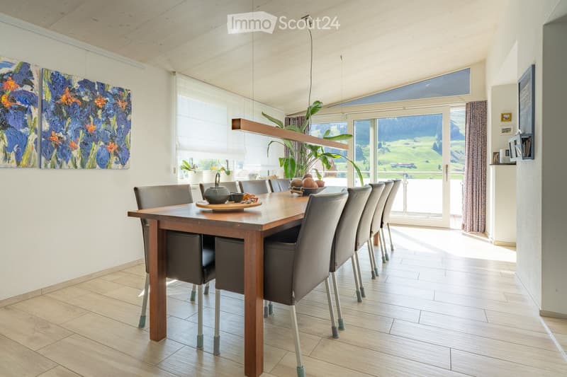 Bright dining room with a wooden table, six chairs, a teapot, and a bowl on the table. Large windows offer a scenic view of a green landscape. The room features modern decor, including paintings and a plant.
