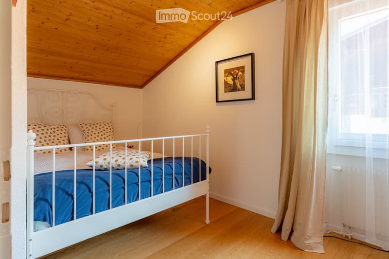Cozy bedroom with a white metal bed, blue blanket, patterned pillows, and a framed picture on the wall. The room features wooden flooring and a slanted wooden ceiling.