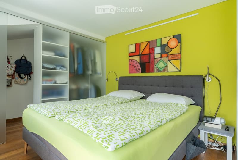 Bright bedroom with a double bed, green and white bedding, a nightstand with a lamp, a colorful abstract painting on the wall, and a modern closet with glass doors.
