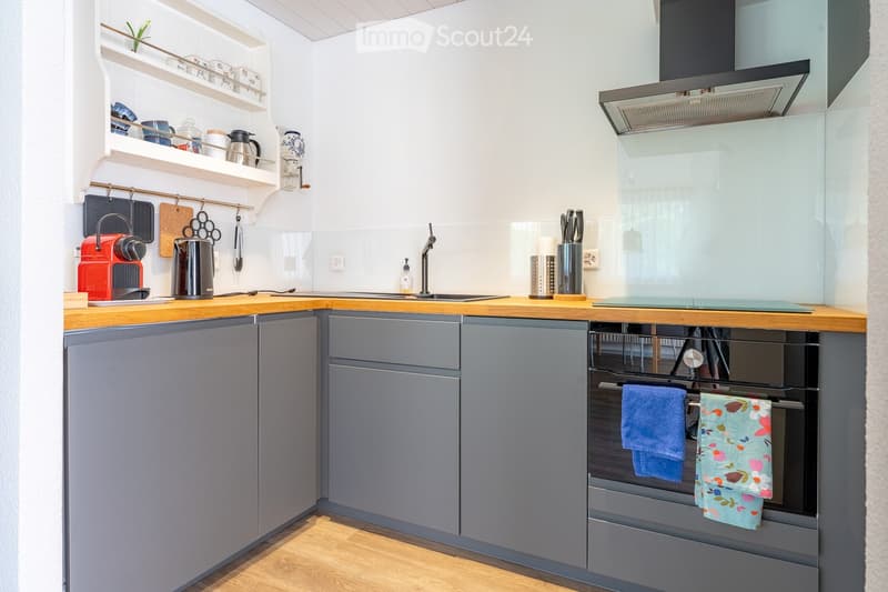 Modern kitchen with grey cabinets, wooden countertops, stainless steel appliances, and a built-in oven. Shelves hold cups and kitchen items, and a knife block is visible on the counter.