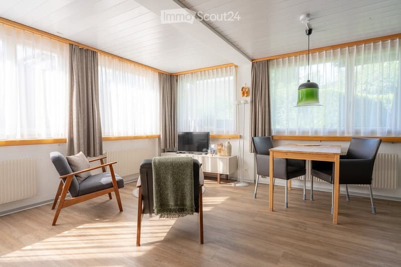 Bright living room with wooden flooring, large windows with curtains, a sofa, armchair, coffee table, dining table with chairs, and a television on a stand.