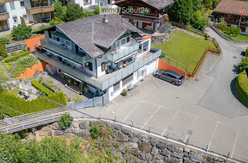 Aerial view of a modern two-story house with a grey roof, large windows, and a spacious balcony. The house is surrounded by a well-maintained garden and has a driveway with a parked car.