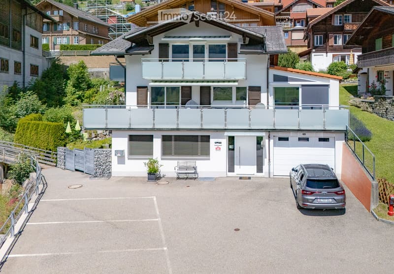 A modern two-story house with a large balcony, garage, and parking space. The house features large windows, a sleek design, and is surrounded by greenery and neighboring houses.