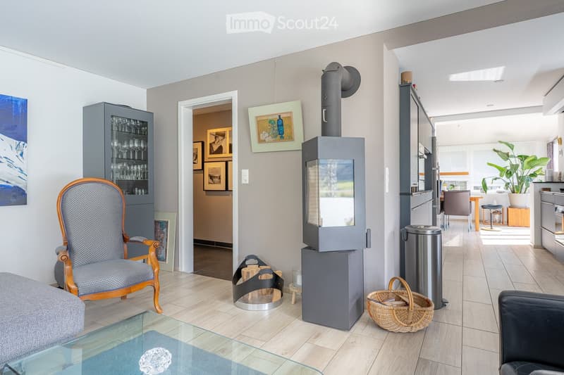 Modern living room with a fireplace, a glass coffee table, a grey sofa, a wooden armchair, a cabinet with glassware, and a basket on the floor.