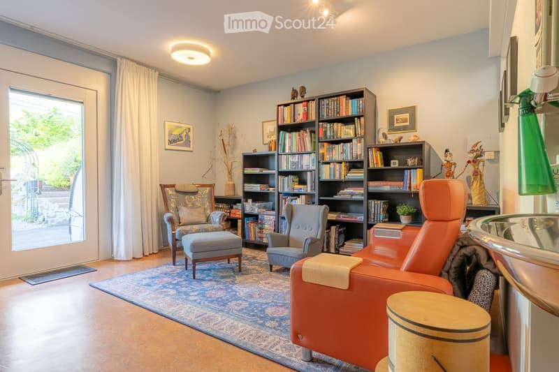 A cozy living room with a large bookshelf filled with books, a patterned rug, a wooden chair, a grey armchair, and an orange sofa. The room features large windows with white curtains and decorative items on the shelves.