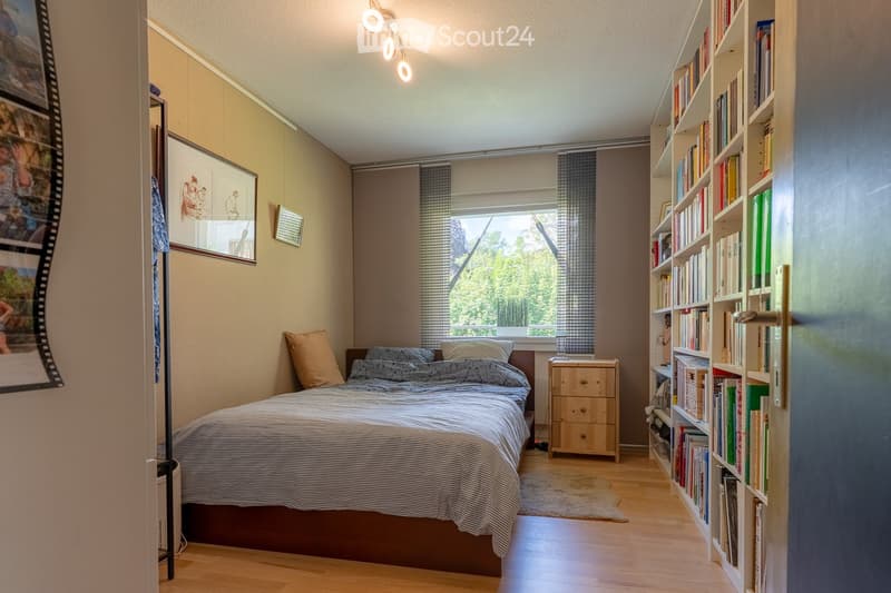 Cozy bedroom with a wooden bed, nightstand, bookshelf filled with books, and a large window with curtains. The room features light-colored walls and wooden flooring.