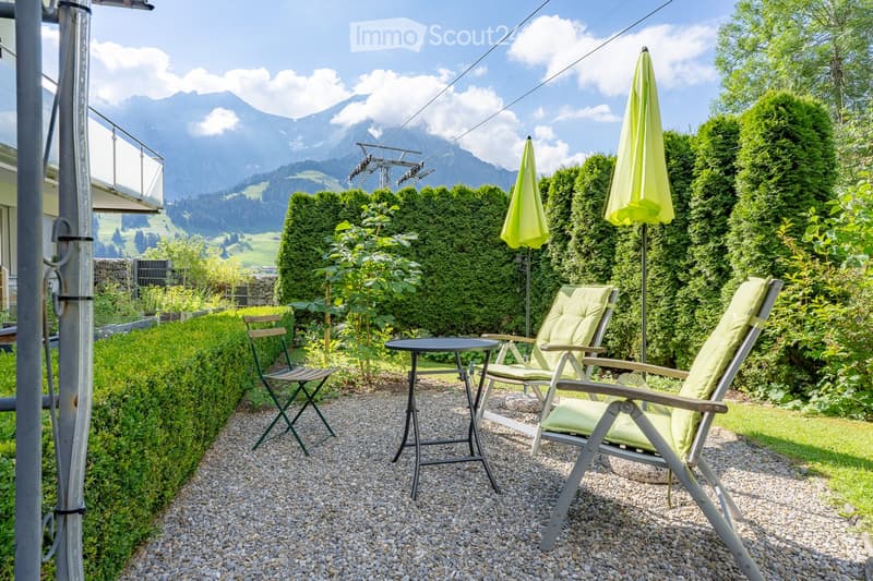A well-maintained garden area with gravel ground, featuring a small round table, two lounge chairs, and two umbrellas. The garden is bordered by neatly trimmed hedges and lush greenery, with a scenic mountain view in the background.