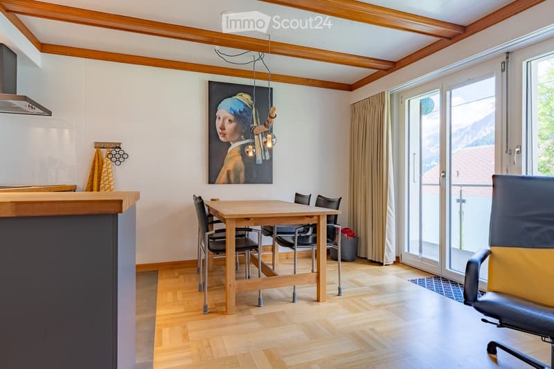 Bright, modern dining area with wooden flooring, a central dining table surrounded by chairs, a sofa, a painting on the wall, and a kitchen island on the left. Large windows and sliding doors lead to a balcony with a scenic mountain view.