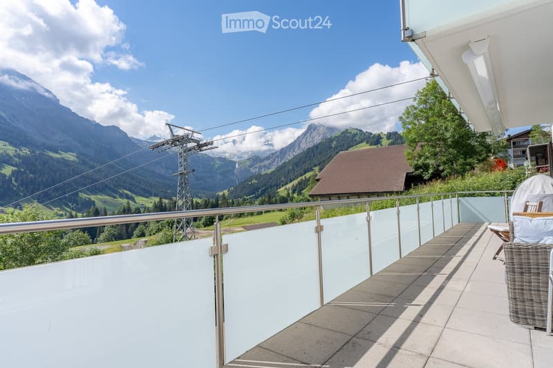 Balcony with a modern glass railing, overlooking a scenic mountain landscape. Includes a wicker chair and a small table.