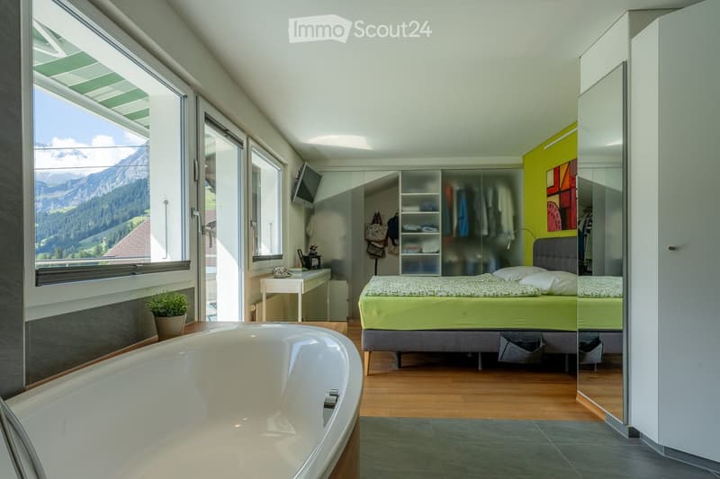 Bright bedroom with a large window offering a mountain view, featuring a bed, bathtub, small table, chair, mirror, wardrobe, and potted plant.