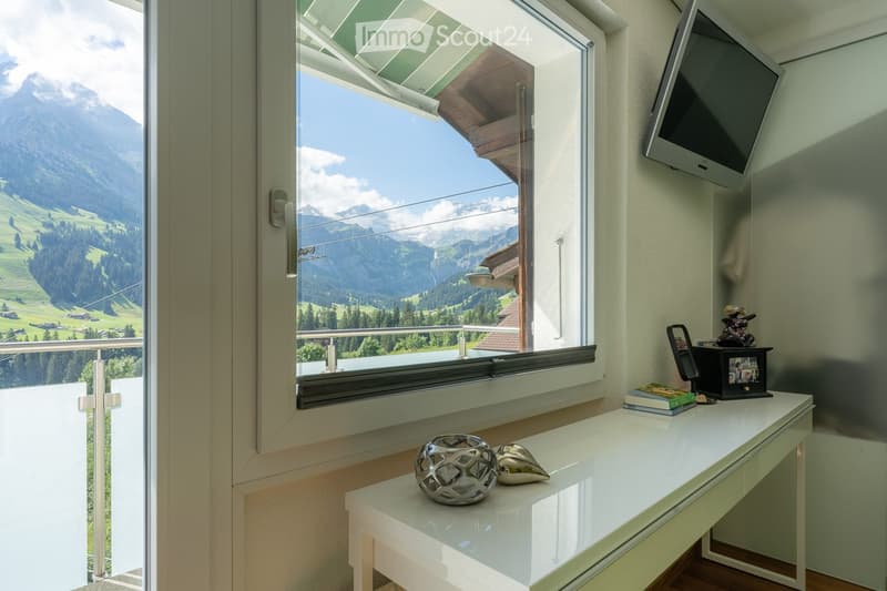 A well-lit room with a large window offering a scenic mountain view, a white table with decorative items, a television mounted on the wall, and a mirror on the table.