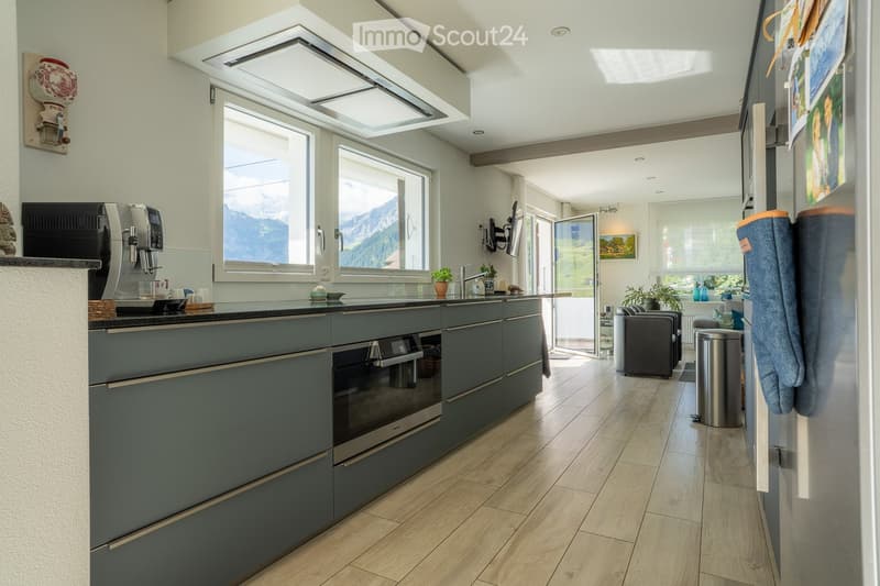 Modern kitchen with grey cabinets, wooden flooring, large windows overlooking mountains, coffee machine, oven, and sink.