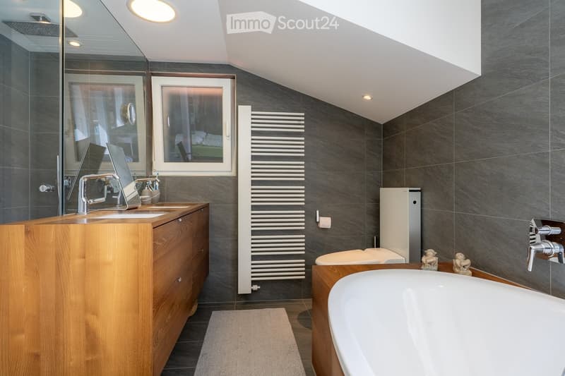 Modern bathroom with dark grey tiles, wooden vanity, bathtub, radiator, and large window offering a view of the bedroom.