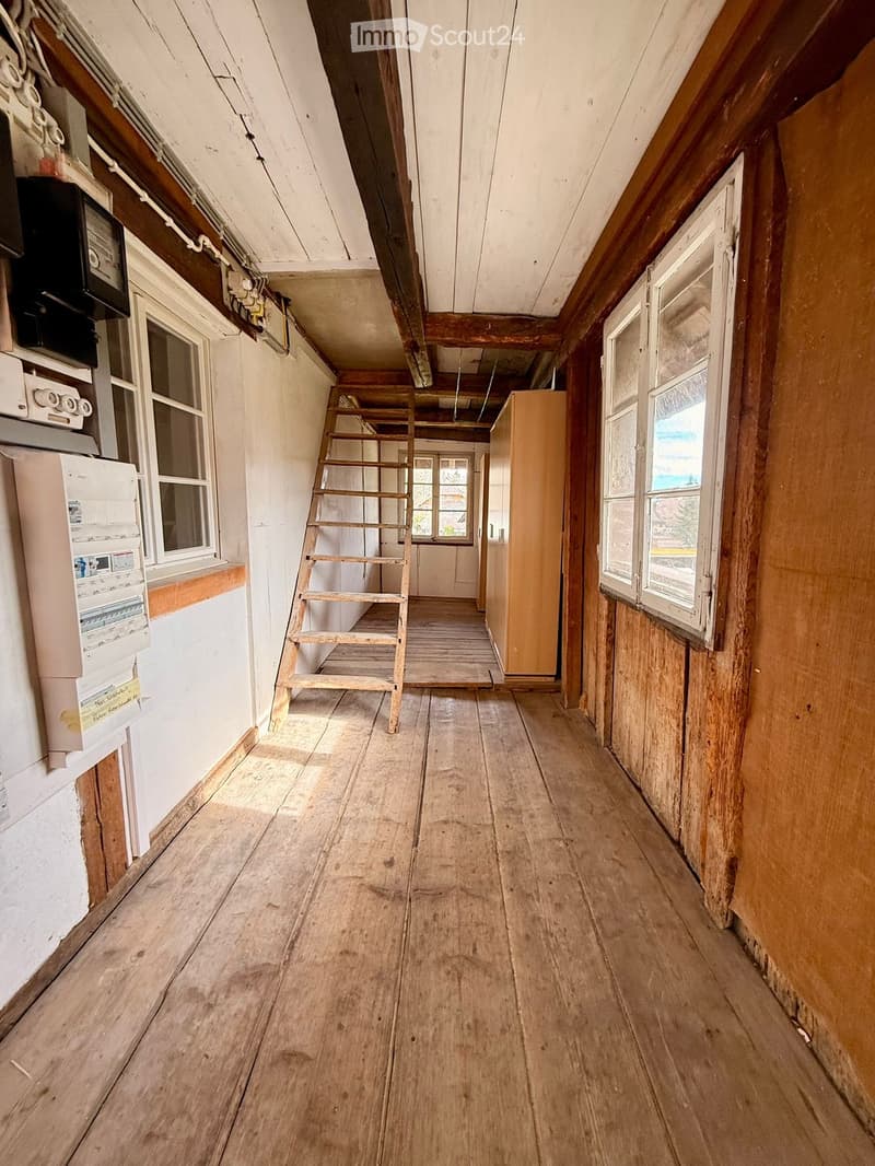 Empty wooden hallway with exposed beams, wooden floor, and a small wooden ladder leading to an upper level. Windows on both sides allow natural light to enter.