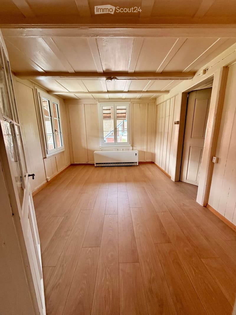 Empty room with wooden flooring, white walls, and a radiator. Two windows and two doors are visible.
