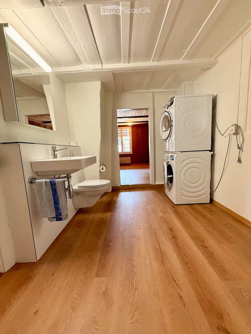 Bathroom with wooden flooring, white walls, sink, toilet, and a stacked washing machine and dryer unit.