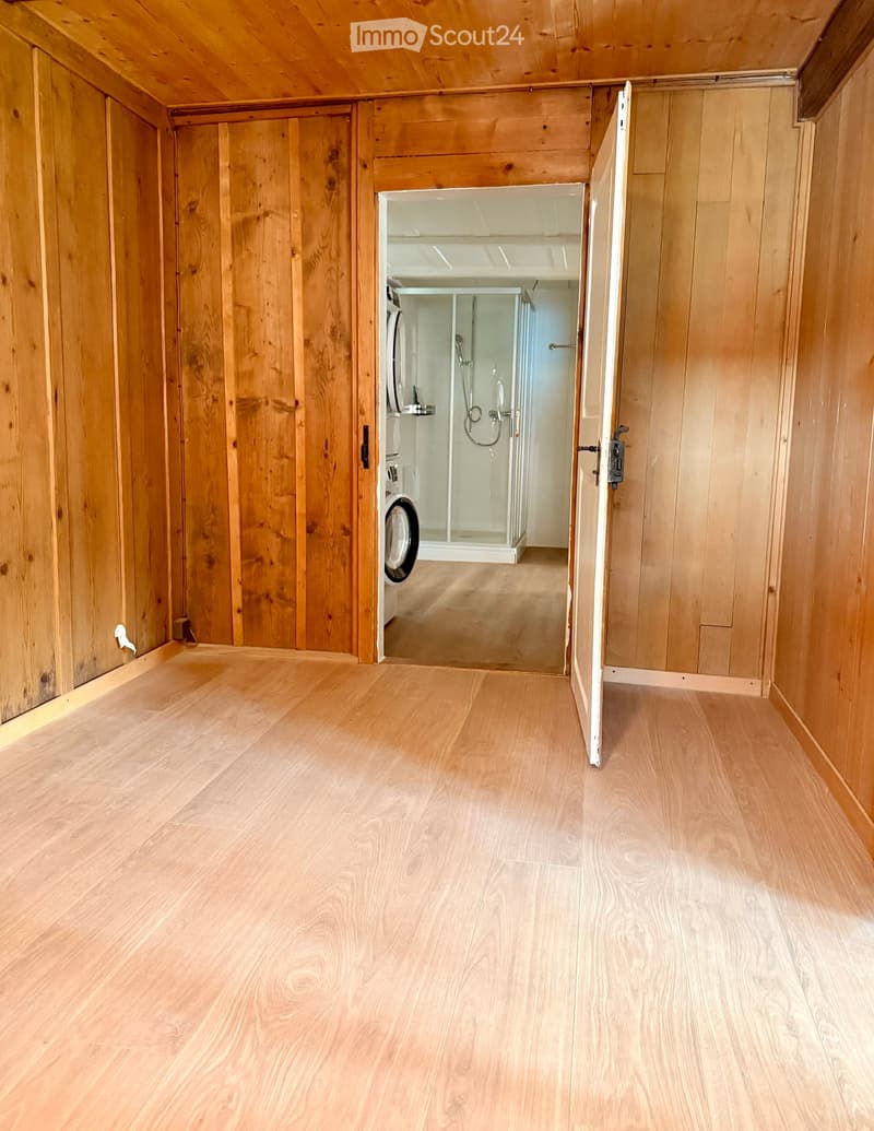 A narrow wooden hallway with light wooden flooring and walls, leading to a private washing machine area and a shower stall.