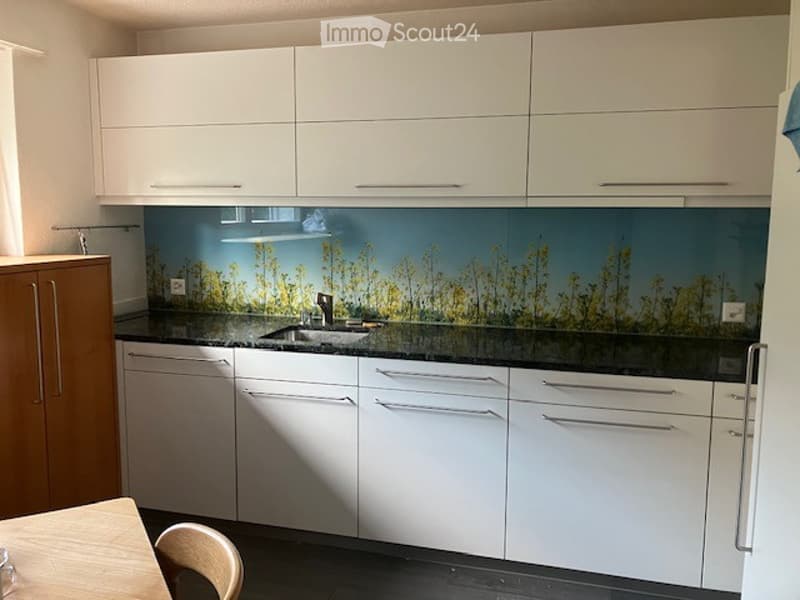 Modern kitchen with white cabinets, black countertops, and a decorative backsplash featuring a nature scene. A wooden chair is visible near the counter.