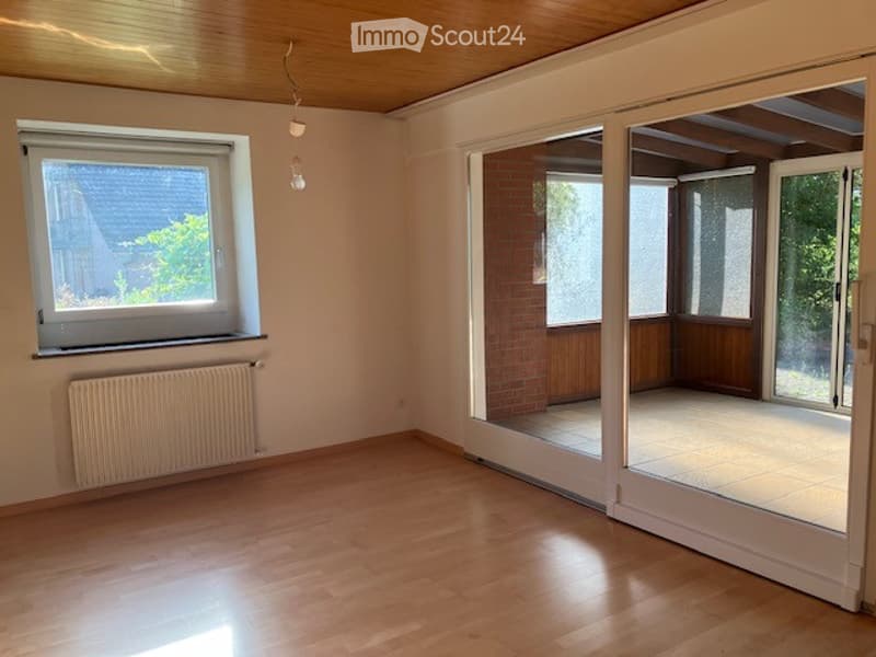 Empty room with wooden flooring, large window with white frame, radiator below, and sliding glass doors leading to a balcony area.