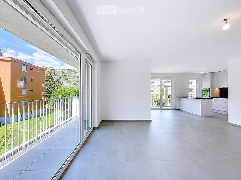 Bright, open living space with large windows and a balcony overlooking a scenic view of greenery and buildings. The room features a tiled floor and white walls, with a modern kitchen area visible in the background.