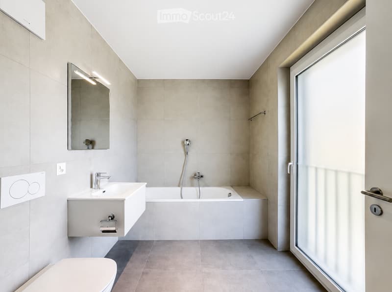 Modern bathroom with a white sink, bathtub, toilet, and a large mirror with built-in lighting. The space features light-colored tiles and a sleek, contemporary design.
