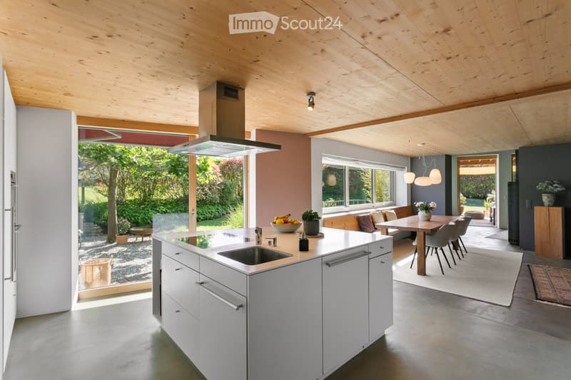 Modern kitchen with white cabinets, island, sink, and dining area. Features pendant lights, a bench, and a view of the garden through large windows.