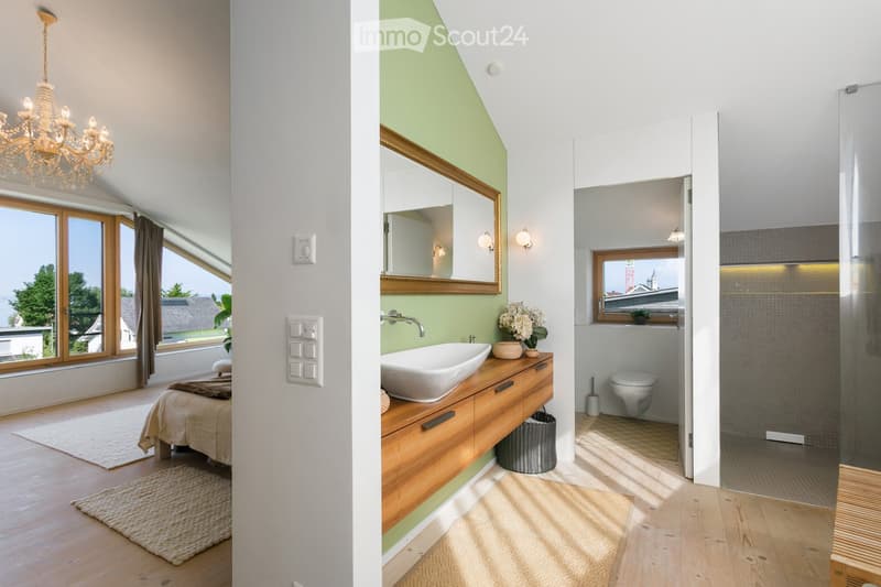 The image shows a modern, well-lit bedroom with large windows and a chandelier, connected to a contemporary bathroom featuring a sink, mirror, toilet, and shower. The bathroom has a sleek wooden cabinet and a vase with flowers on the counter.
