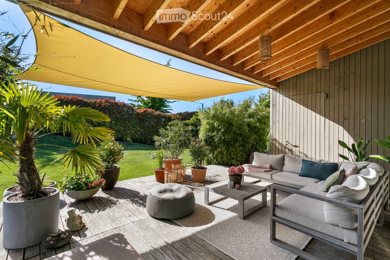 A modern outdoor patio with a wooden deck, covered by a pergola with a retractable awning. Features a grey sectional sofa, a coffee table, an ottoman, and several potted plants, including a large palm tree. The space is surrounded by a well-maintained gar