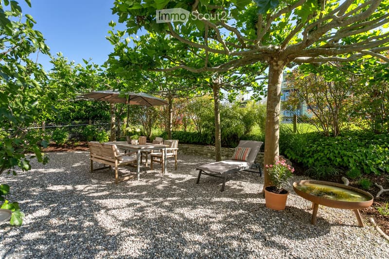 A serene garden area with a gravel-covered ground, featuring a wooden bench, a lounge chair, a small table with chairs, a potted plant, and a decorative water fountain. The area is shaded by large trees and surrounded by lush greenery.