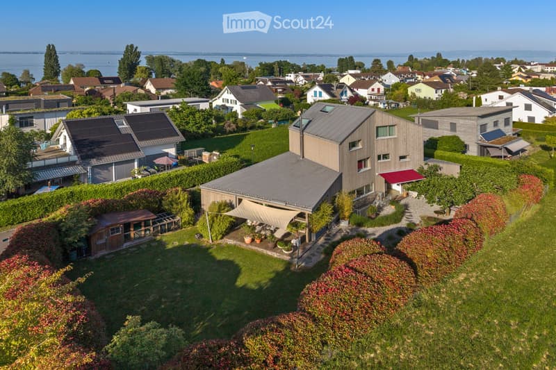 Aerial view of a modern house with a large garden, surrounded by other houses and greenery, with a view of a lake in the background.