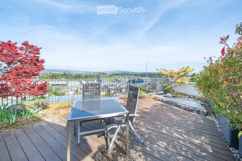 Wooden deck with a glass-top table and chairs, surrounded by plants and a cityscape view.