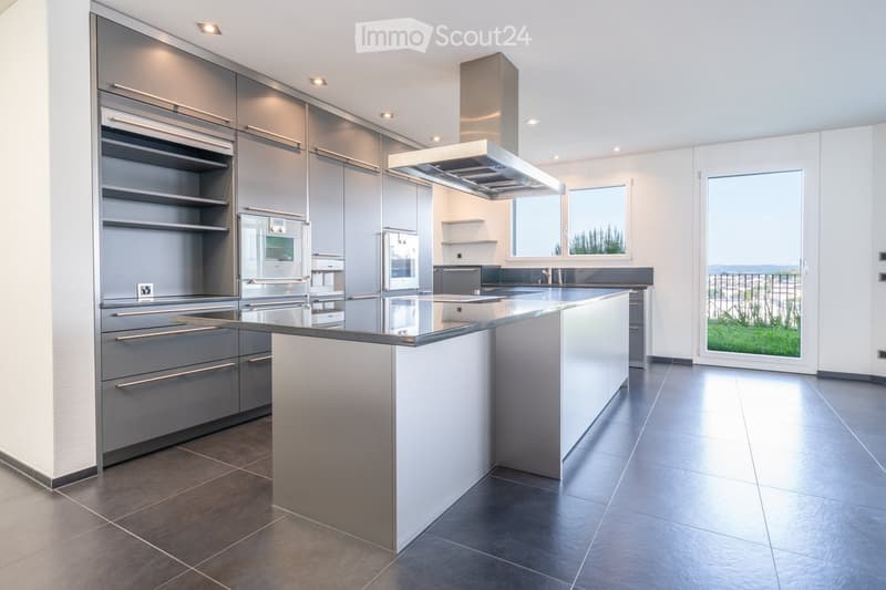 Modern kitchen with stainless steel appliances, a central island, and large windows offering a view of the outdoor area.