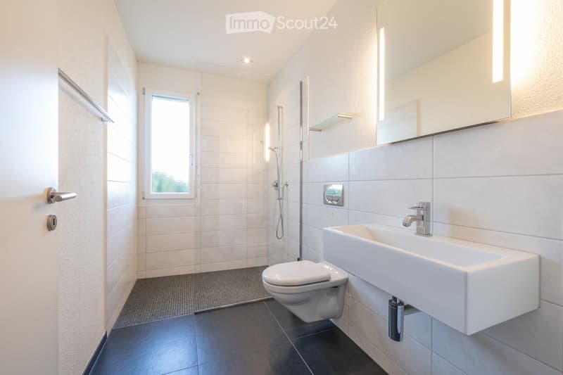 Modern bathroom with white tiles, a large mirror, a sink, a toilet, and a glass-enclosed shower area.