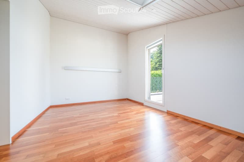 Empty room with light-colored walls, wooden flooring, and a large window with a view of greenery outside.