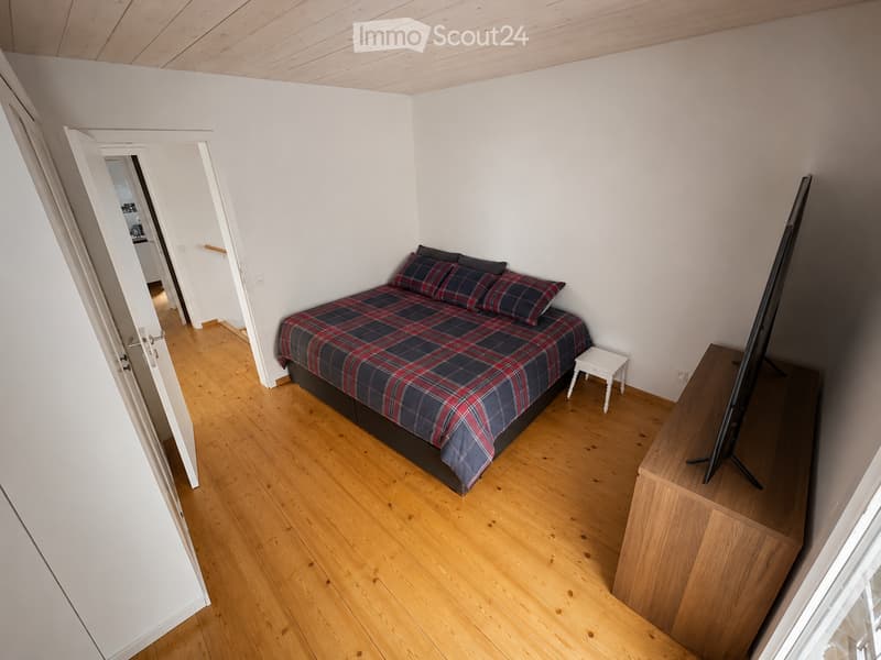 Cozy bedroom with wooden flooring, a neatly made bed with plaid bedding, a small nightstand, and a television on a wooden stand.