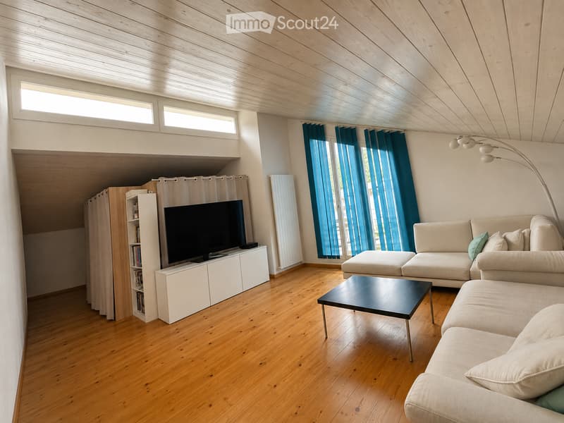 Modern living room with wooden floors, white walls, and large windows with blue curtains. Features a beige sectional sofa, black coffee table, flat-screen TV on a white stand, bookshelf, and a floor lamp.
