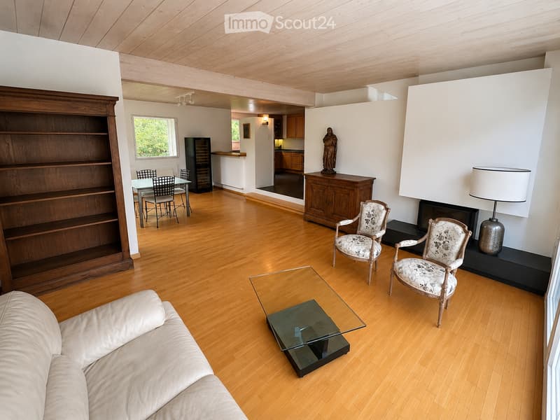Spacious living room with wooden flooring, a cream-colored sofa, glass coffee table, two ornate armchairs, a wooden cabinet, a fireplace with a lamp, and a dining area with a table and chairs.