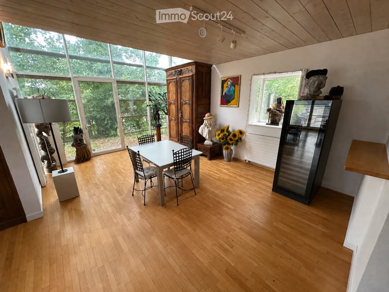 Bright room with wooden flooring, large windows, a dining table with chairs, a cabinet, a vase with sunflowers, a painting, and sculptures.