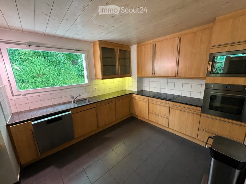 Modern kitchen with wooden cabinets, stainless steel appliances, and a large window overlooking greenery.