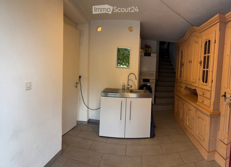 A compact kitchen with a sink, faucet, white cabinets, a washing machine, and a wooden cupboard with glass doors.