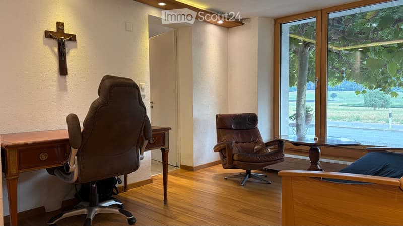A well-lit room with wooden flooring, a desk with a chair, a small table with a leather chair, and large windows offering a view of greenery outside.