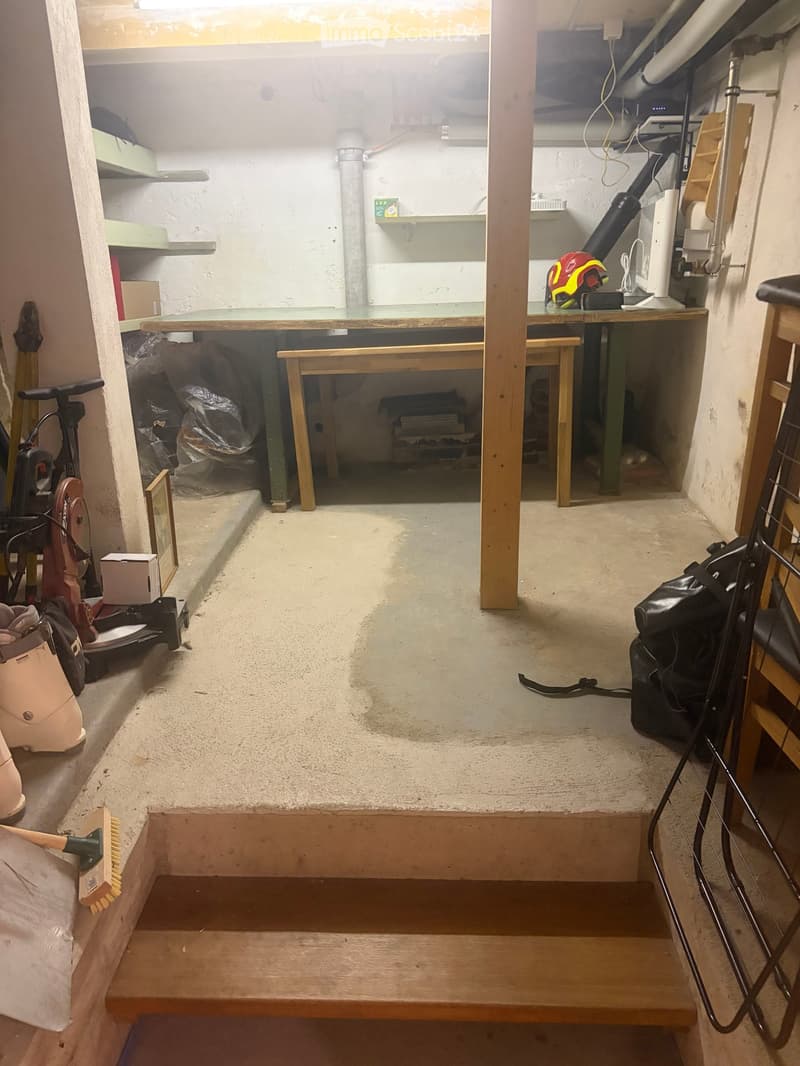 A basement or utility room with a concrete floor, a wooden table and chairs, shelves, a backpack, a helmet, and various tools and equipment.