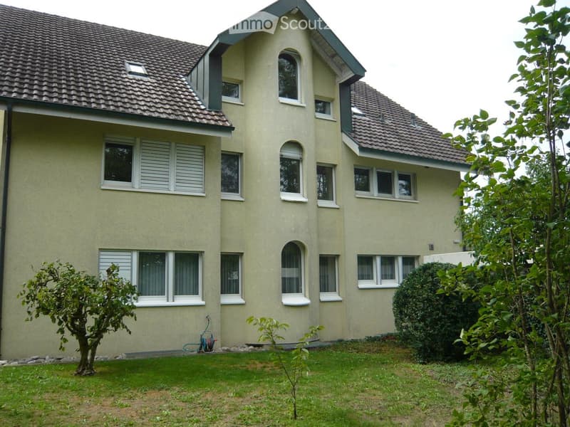 Two-story house with beige siding, multiple windows, and a well-maintained lawn with small trees and shrubs.
