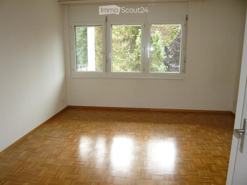 Empty room with wooden parquet flooring and large windows showing greenery outside. No furniture or equipment present.