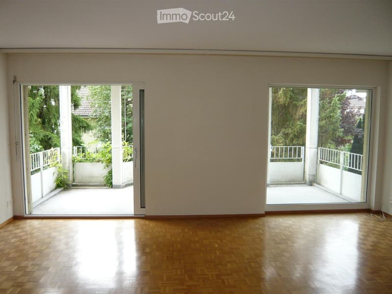 Empty room with wooden flooring and large sliding glass doors leading to a balcony area with greenery visible outside.