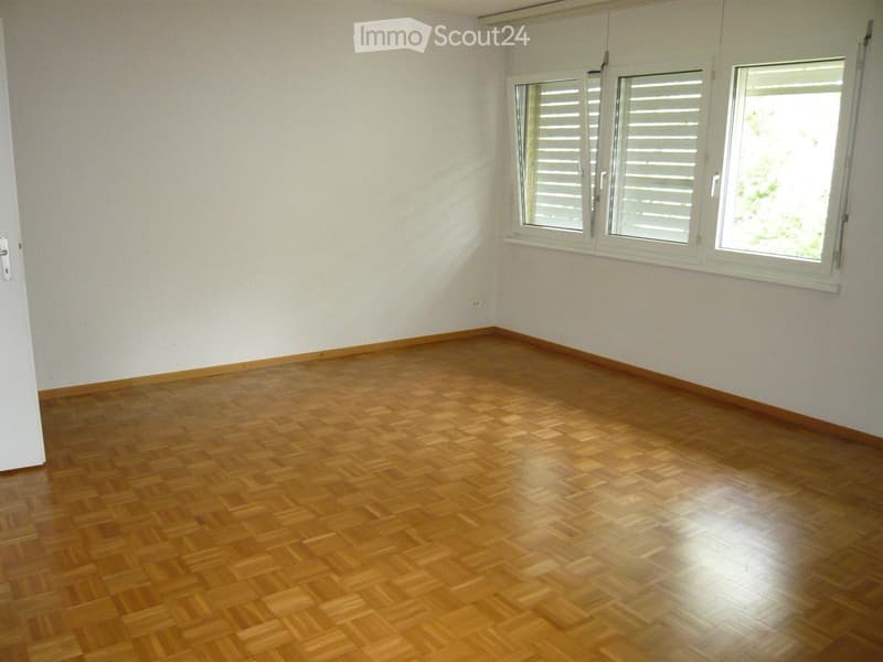 Empty room with wooden flooring and white walls, featuring two windows with shutters and a door on the left side.
