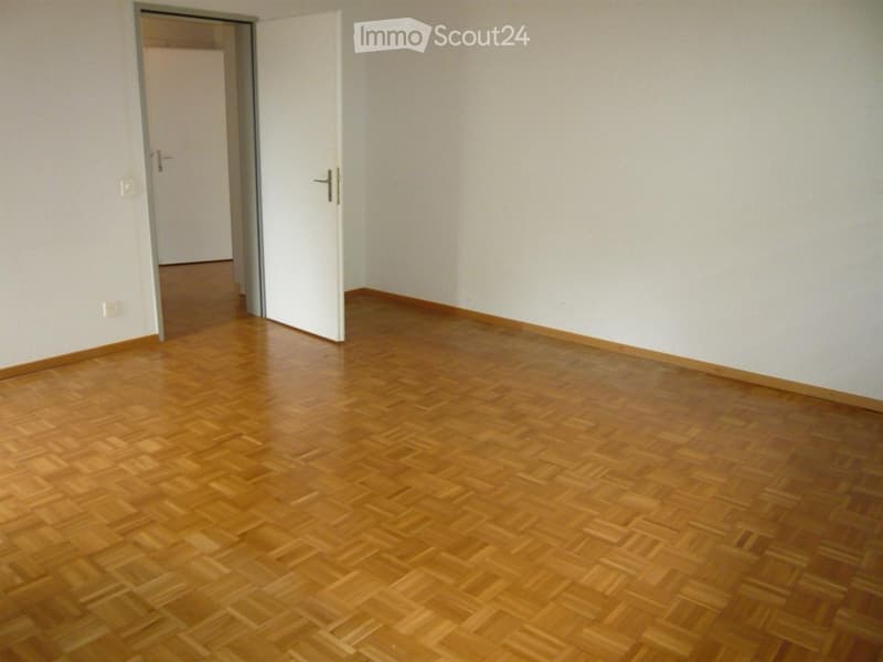 Empty room with wooden flooring and white walls, featuring a partially open white door leading to another space.
