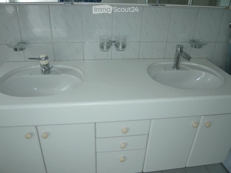 Modern bathroom with a white double sink, chrome faucets, and two glass cups on the countertop. White cabinets with round knobs are below the sinks.