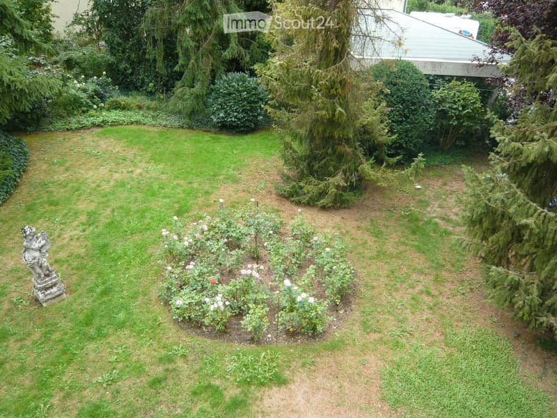 Well-maintained garden with a lawn, rose bushes, and a small statue on a pedestal. The garden is surrounded by trees and shrubs, with a house partially visible in the background.
